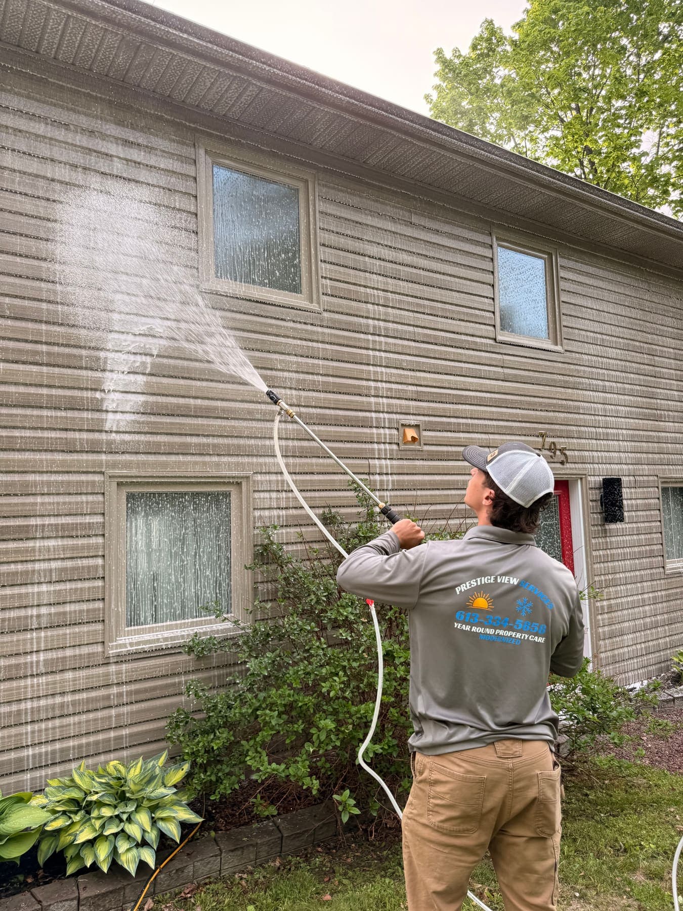 PVS crew member soft-washing tan vertical siding on a residential property.